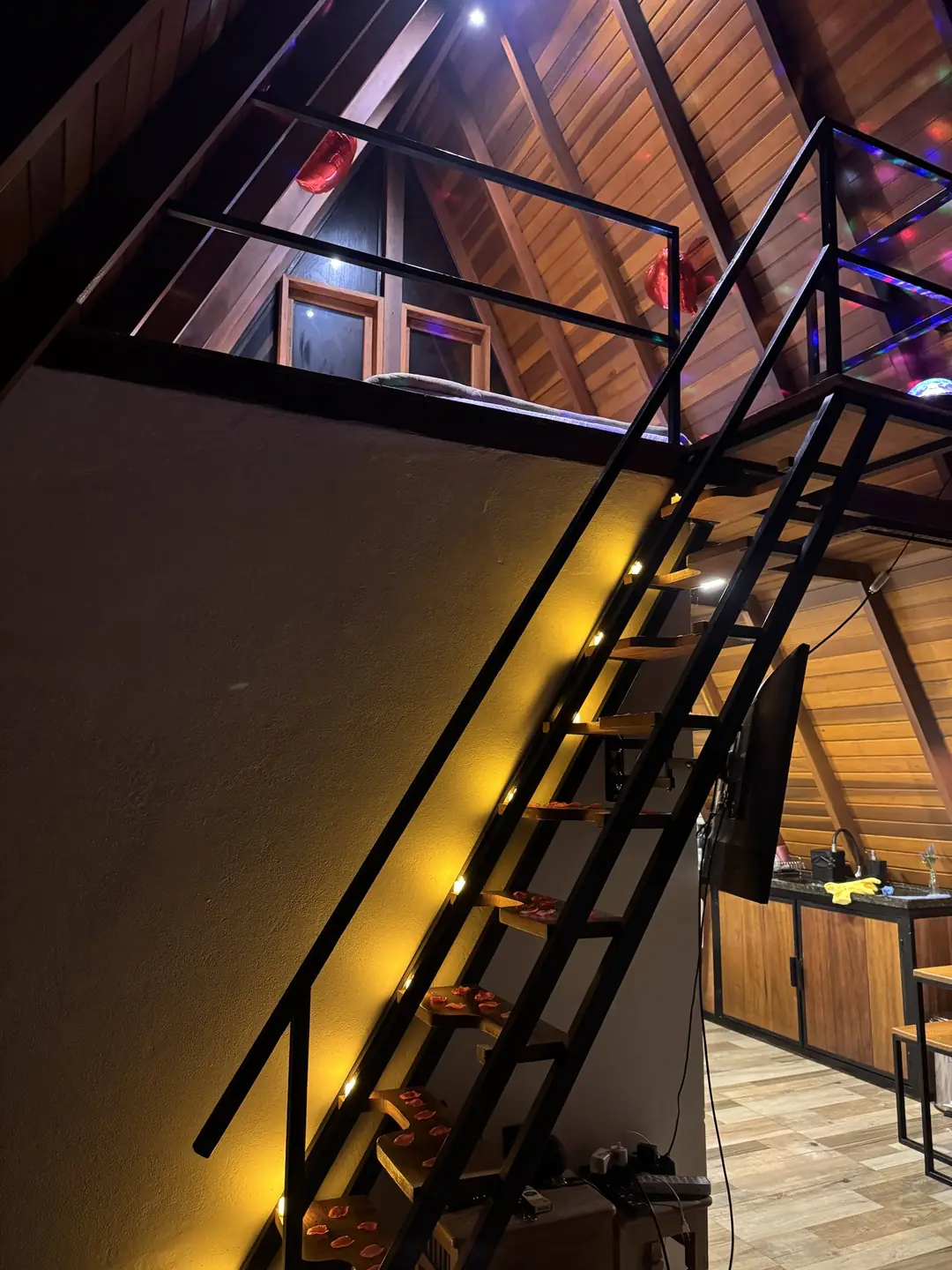 Mezanino da cabana A-frame visto de baixo, com as luzes coloridas do globo giratório espalhadas pela madeira