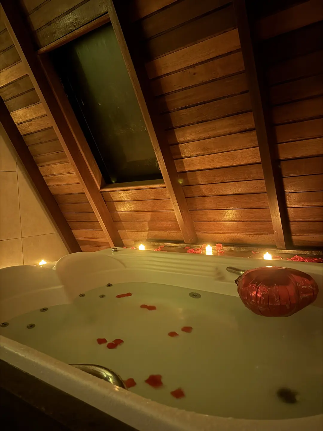 Hidromassagem cheia com pétalas vermelhas na água, velas de LED acesas na borda e um balão de coração apoiado